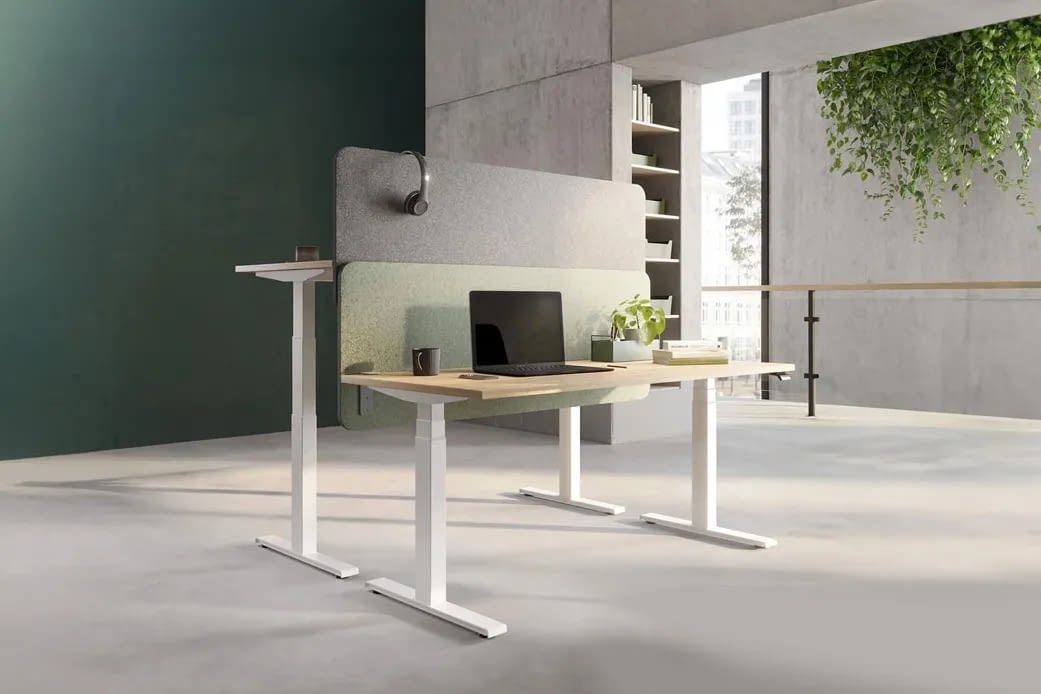 Doppelarbeitsplatz mit zwei elektrisch höhenverstellbaren Schreibtischen in Weiß mit Holzdekorplatte, einer auf Sitzhöhe und einer auf Stehhöhe. An den Stirnseiten der Tischplatten montierte Akustiktrennwände in Grau und Grün dienen als Sichtschutz und strukturieren die Arbeitsplätze.