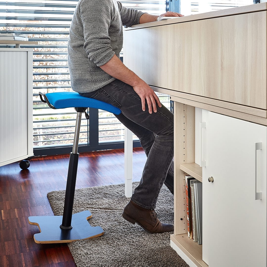 Seitenansicht vom Stehhocker sella activa in blau mit einer Person beim Arbeiten am Arbeitsplatz im Büro.