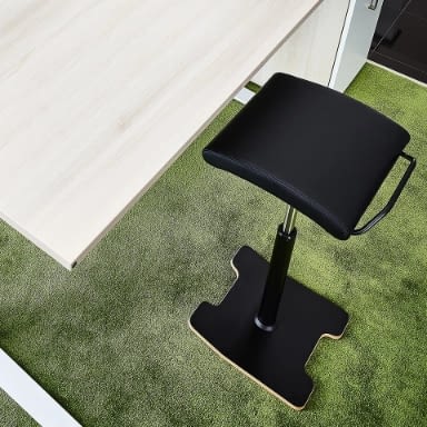 Der Stehhocker sella activa in schwarzem Echtleder ist in der Draufsicht neben einem Schreibtisch im Büro positioniert.