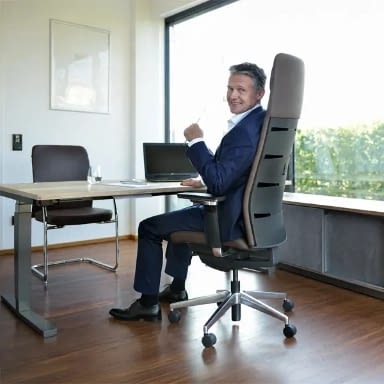 Eine Person sitzt lächelnd am Schreibtisch auf dem Chefsessel agilis matrix aus echtem Leder mit Kopfstütze und Nackenkissen, im Hintergrund ergänzt ein Besucherstuhl die Büroszene.