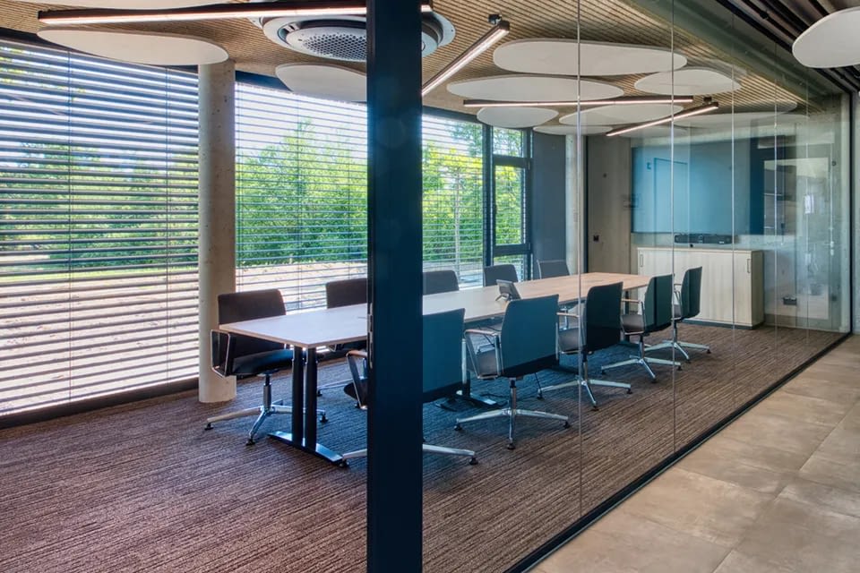Im modernen Meetingraum stehen ergonomische Drehstühle mit Armlehnen an einem langen Konferenztisch aus Schreibtischen, ergänzt durch Glasfront und große Fensterflächen.
