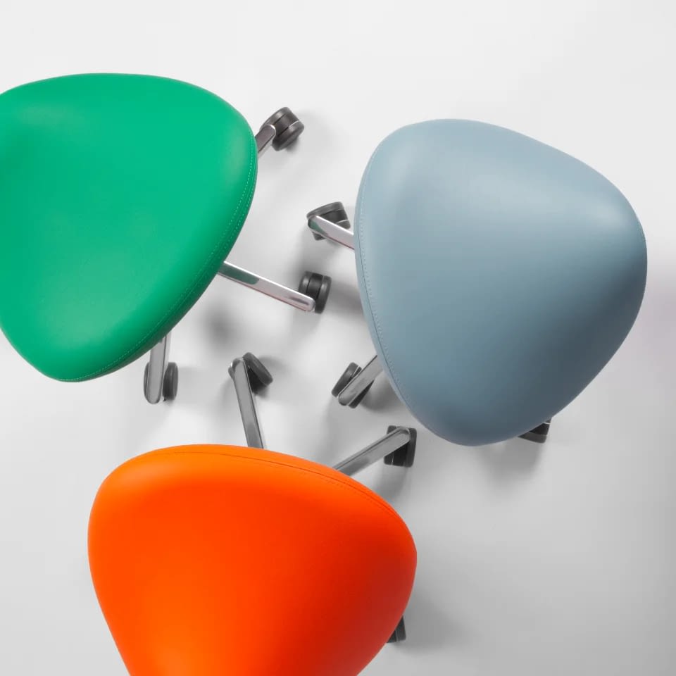 Die Sattelhocker sella sind als Gruppe in Blau, Grün und Orange auf hellem Studiohintergrund dargestellt und zeigen die Varianten im Kunstlederbezug.