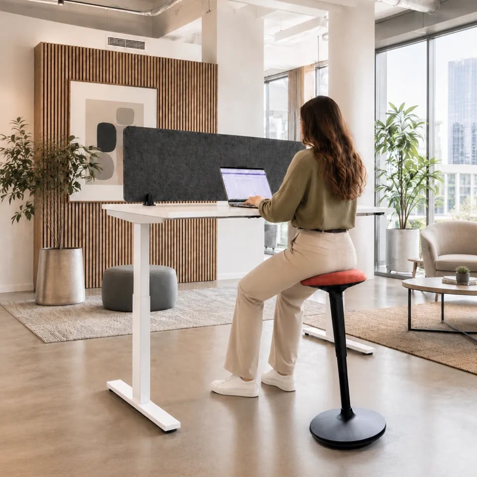 In einem Büro mit warmer Farbgebung arbeitet eine Person am Schreibtisch auf dem Stehhocker sella flex in Korallenrot und sitzt dabei in dynamischer Bewegung.
