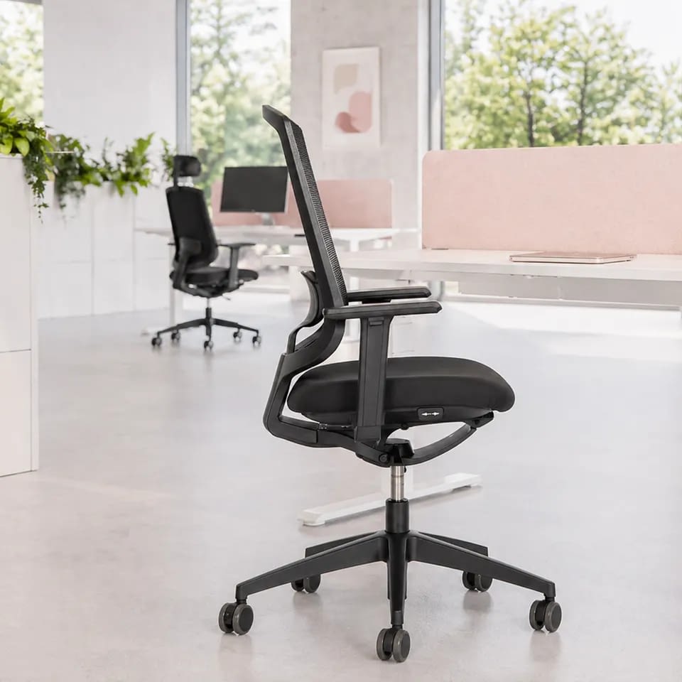 Im hellen Büro ist der ergonomische Bürostuhl laboro in Schwarz in seitlicher Ansicht an weißen Sitz-Steh-Schreibtischen mit Sichtschutz für eine moderne Arbeitsplatzgestaltung zu sehen.